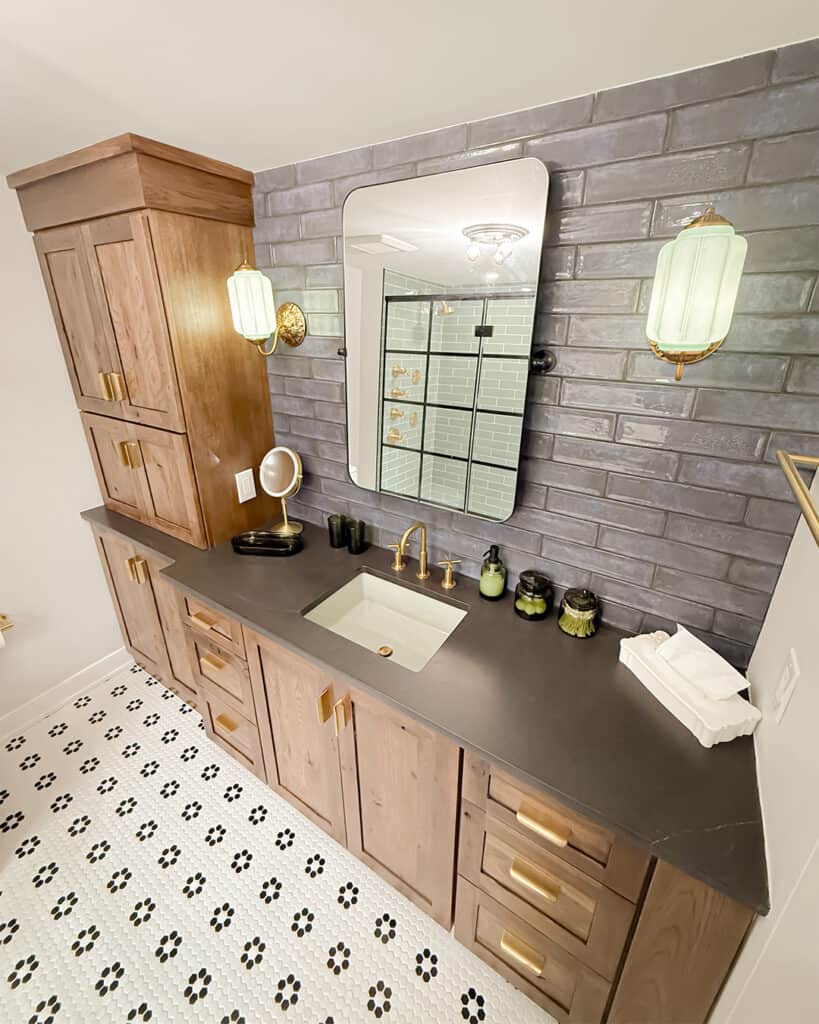 A bathroom vanity with wood cabinets, black countertop, brass fixtures, and a rectangular sink. A large mirror is mounted on a gray tiled wall, flanked by two green glass sconces above a patterned black and white tile floor.