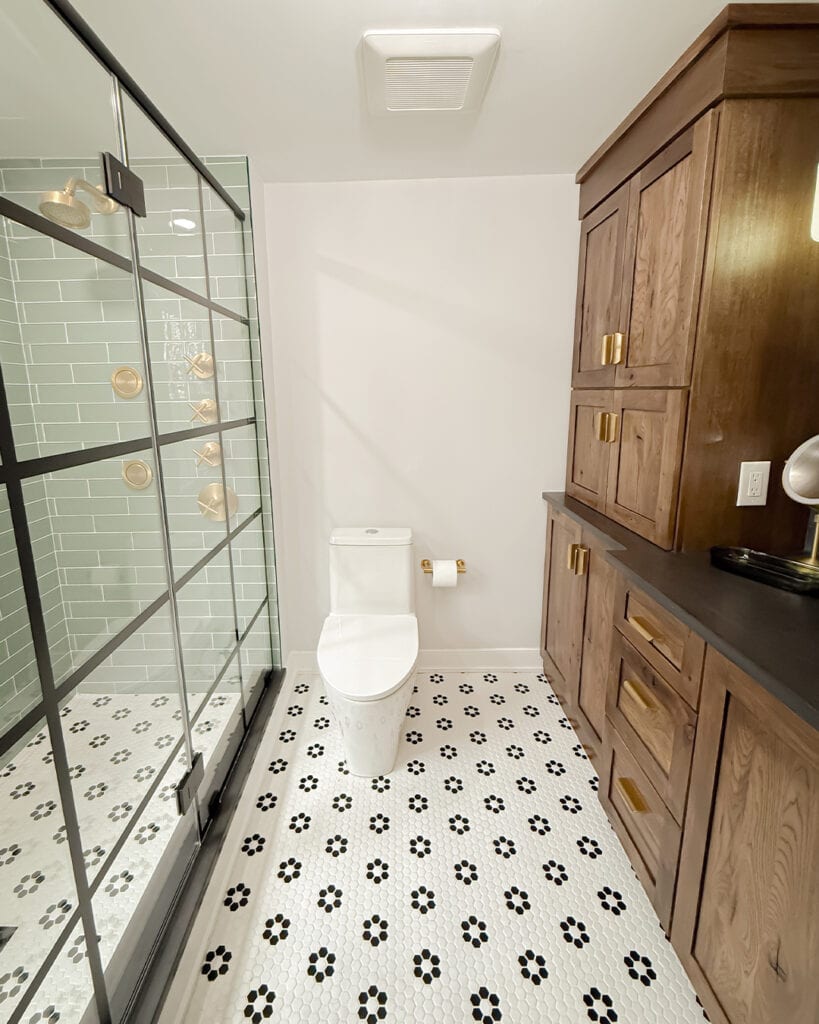 Modern bathroom with a glass-enclosed shower, white toilet, wood cabinetry with gold handles, black countertop, and white floor tiles with black floral patterns. The walls are light-colored, creating a bright look.