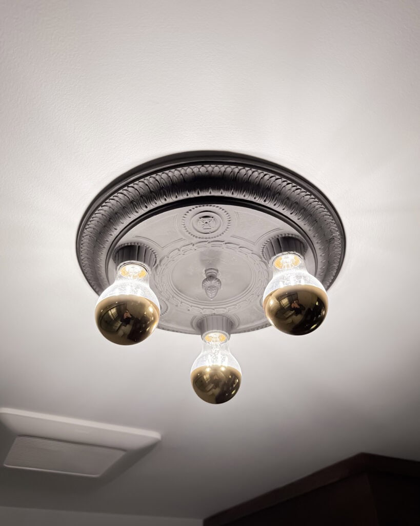 A ceiling light fixture with an ornate, circular black and white base holds three light bulbs; two bulbs have gold reflective bottoms, and one bulb is clear and illuminated. The ceiling is white and smooth.