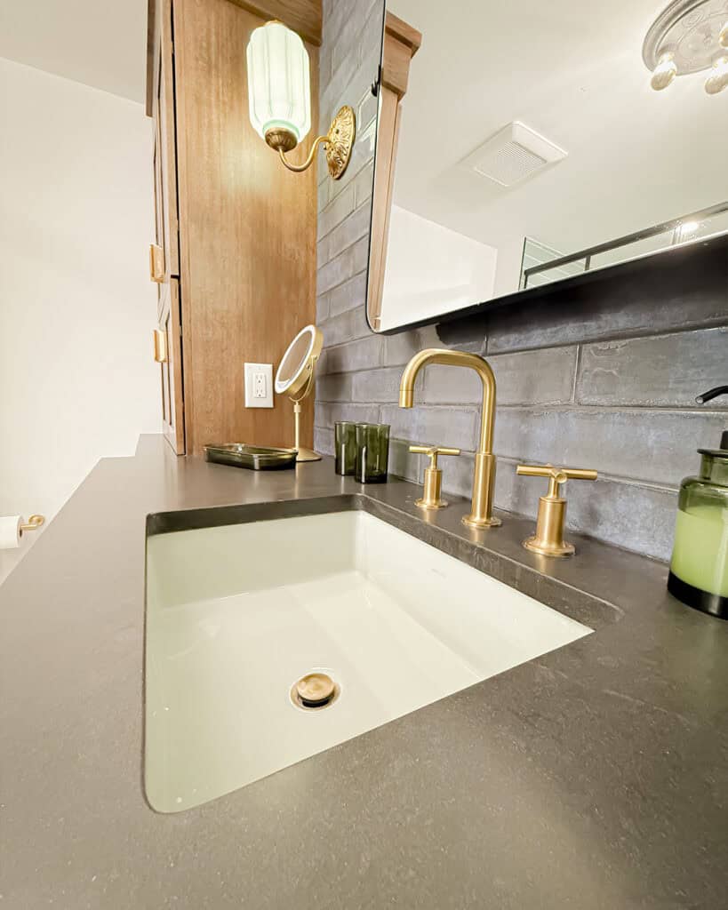 Modern bathroom vanity with a rectangular under-mount sink, matte black countertop, and brushed gold faucet. Behind the sink are green glass accessories, a small mirror, and a wall sconce with a green shade.