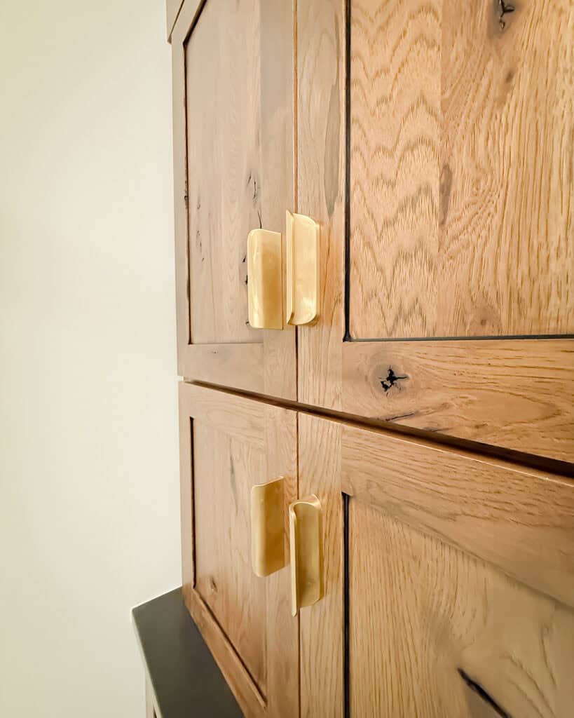 Close-up of wooden cabinet doors with brass handles. The wood has a natural grain and knot details, and the cabinet is set against a plain light-colored wall.
