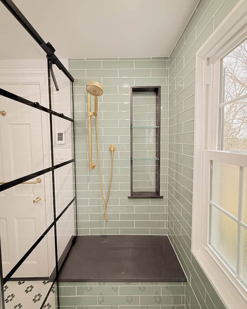 Modern shower with green subway tiles, gold handheld showerhead, black shower bench, built-in black-framed niche, glass partition, and large window letting in natural light. Paw print tiles are visible on the floor.