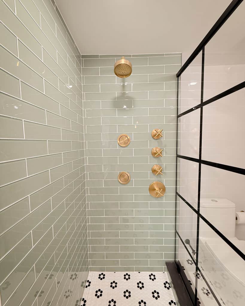 A modern shower with pale green subway tiles, gold fixtures, and a glass partition with black grid lines. The floor has white tiles with small black flower designs. A toilet is visible outside the shower area.