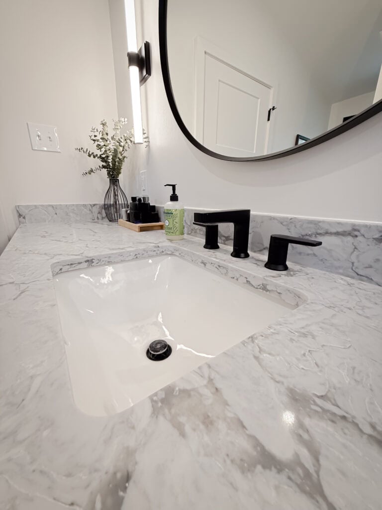A modern bathroom sink with a marble countertop, matte black faucet, liquid soap, and a decorative vase with greenery, set beneath a round mirror and bright wall light.
