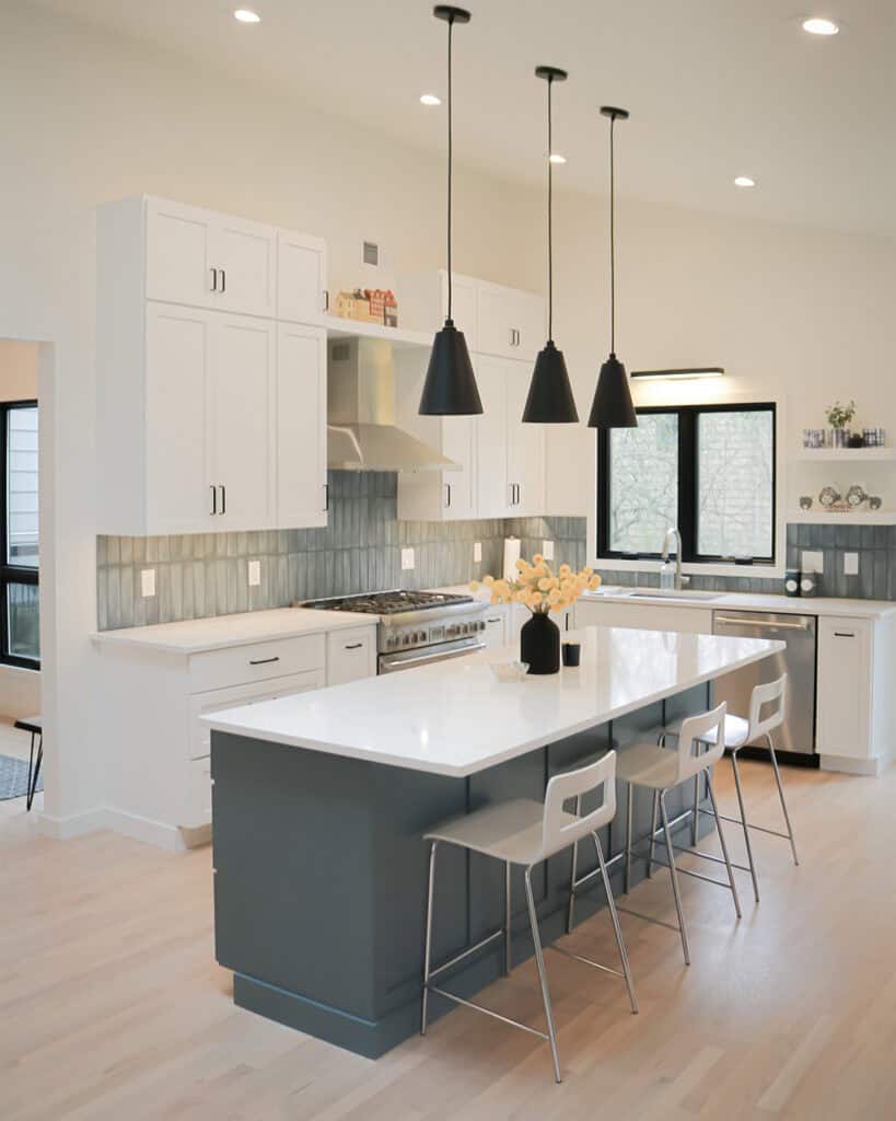 Modern kitchen with white cabinets, a large gray island with three white barstools, three black pendant lights, stainless steel appliances, and a vase of flowers on the island. Light wood flooring and large windows provide natural light.