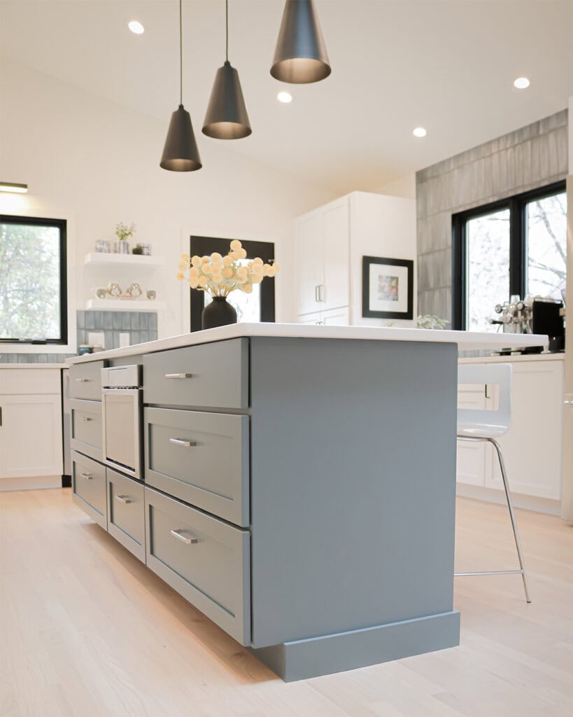 Modern kitchen with a large gray island, sleek drawers, white countertops, pendant lighting, bar stools, and large windows letting in natural light. Minimalist decor with light wood flooring and white cabinetry.