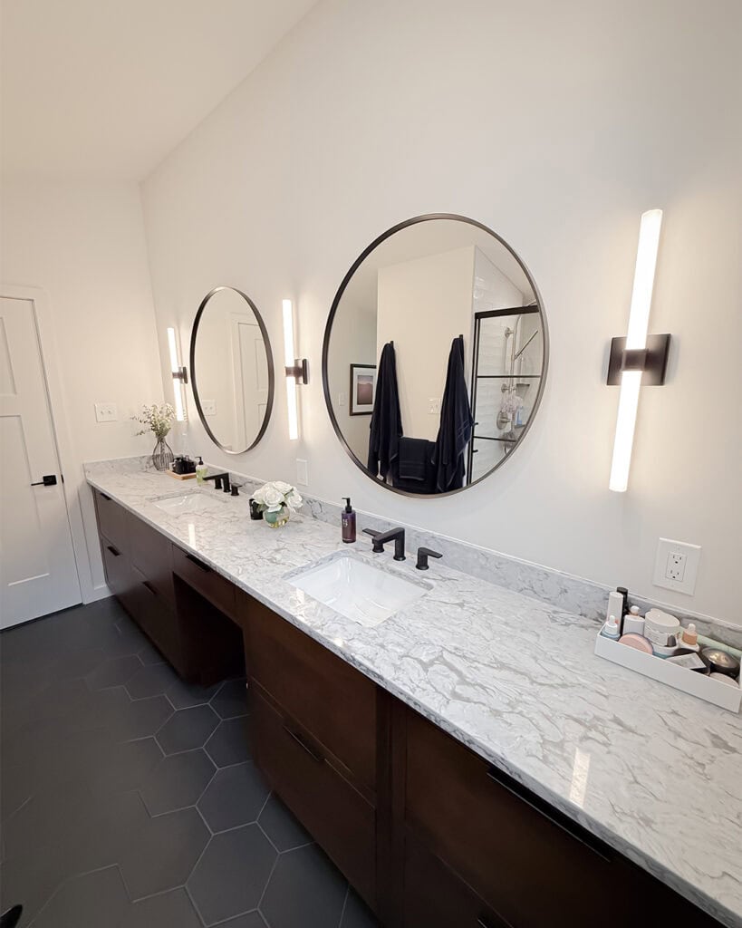 Modern bathroom with two round mirrors, marble countertop, dark wood cabinets, hexagonal floor tiles, wall-mounted lights, and neatly arranged toiletries and flowers on the counter. A shower and towels are visible in the background.