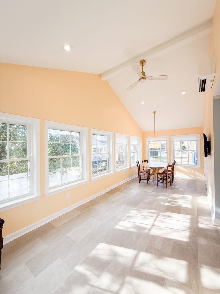 A bright sunroom with peach walls, light tile flooring, large windows along one side, a ceiling fan, and a wooden dining table with four chairs near the far end. Sunlight streams through the windows.