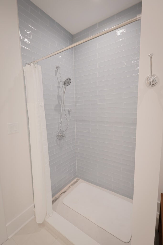 A modern shower with light blue subway tile walls, a white shower curtain partially pulled back, a handheld showerhead, and a white floor mat on a clean shower floor.