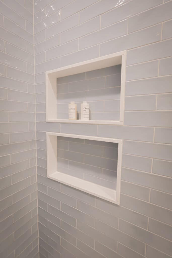 Two white built-in shower shelves set into a light blue subway tile wall, with two bottles of hair or body care products placed on the upper shelf. The lower shelf is empty.