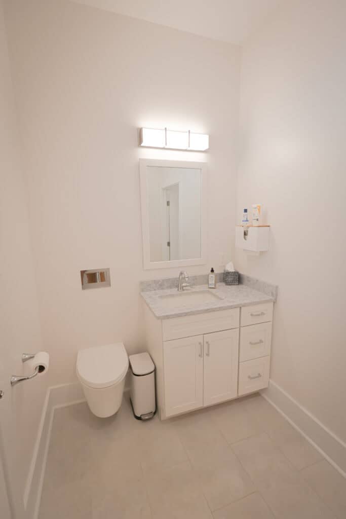 A modern, minimalist bathroom with white walls, a wall-mounted toilet, a small trash bin, a vanity cabinet with a marble countertop, a sink, a mirror, and a wall-mounted light fixture above the mirror.