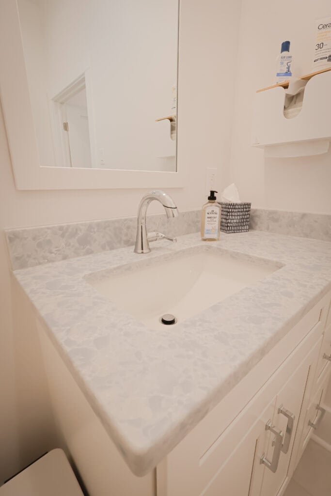 A bathroom vanity with a light-colored, speckled countertop, an undermount sink, a silver faucet, and a mirror above. Soap, tissue box, and skincare products are visible on the counter and on a wall shelf.