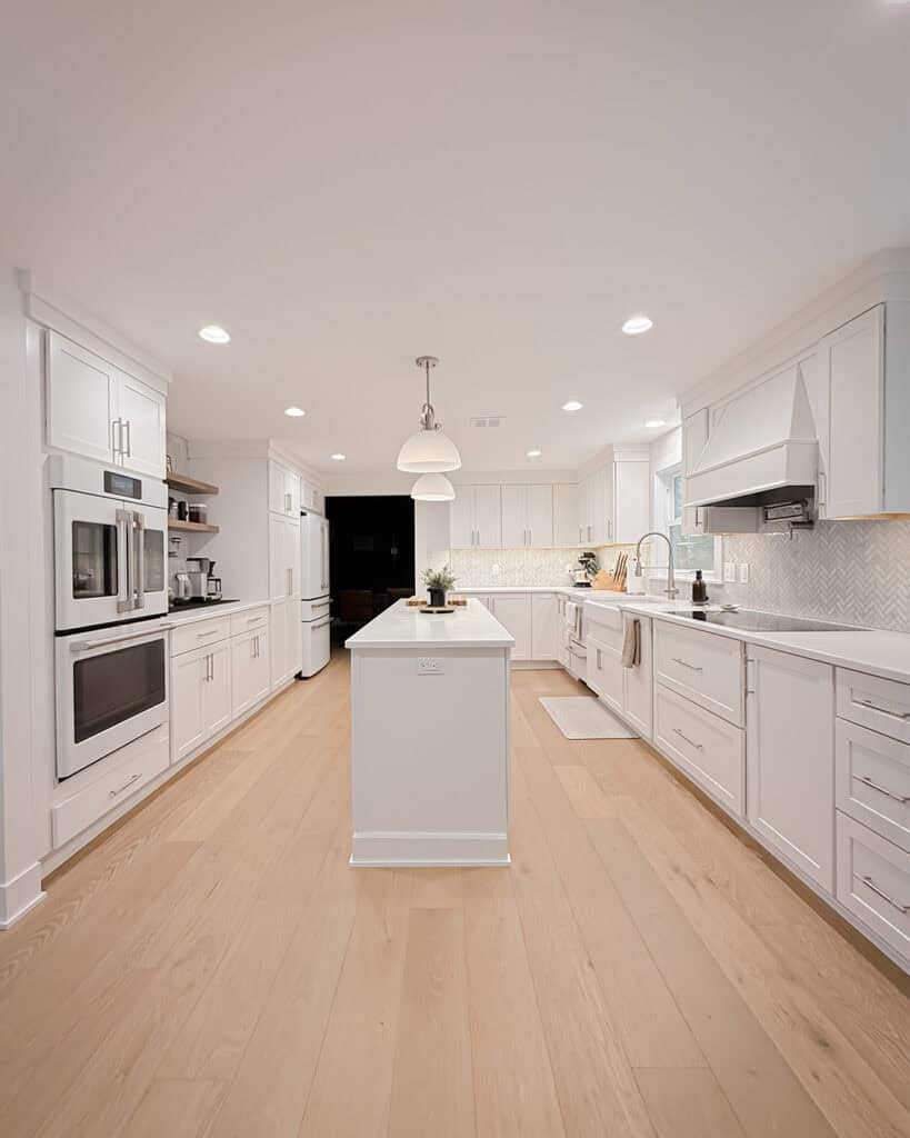 Bright, modern kitchen with white cabinets, stainless steel appliances, light wood floors, a central island with a plant, pendant lighting, and a chevron-patterned backsplash.