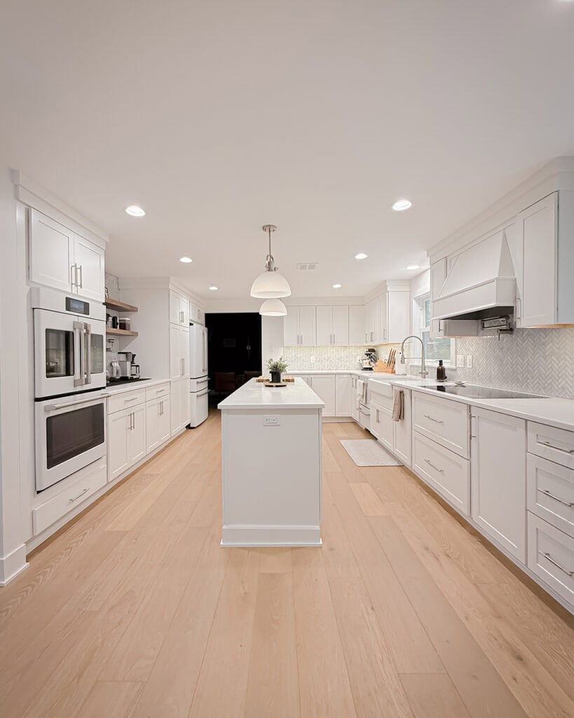 A modern, bright kitchen with white cabinets, stainless steel appliances, light wood flooring, a central island with a plant, and pendant lighting overhead. The space is clean and well-organized.