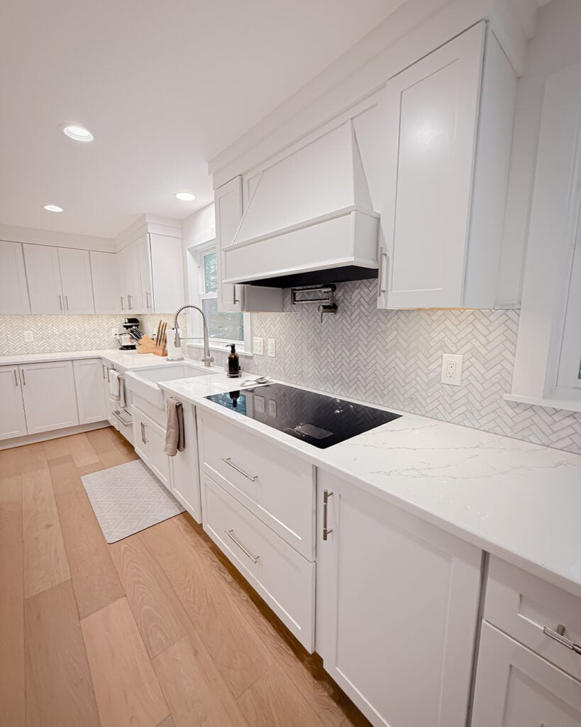 Modern kitchen with white cabinets, marble countertops, light wood floors, a built-in stovetop, farmhouse sink, and herringbone tile backsplash. Kitchen utensils and small appliances are neatly arranged on the counter.