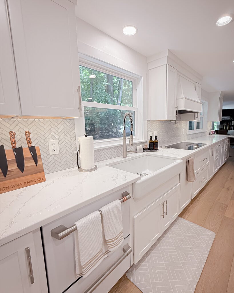 Bright, modern kitchen with white cabinets, marble countertops, a farmhouse sink, paper towel roll, knives on a wooden block, and light wood floors. Large window above the sink and geometric patterned gray rug on the floor.