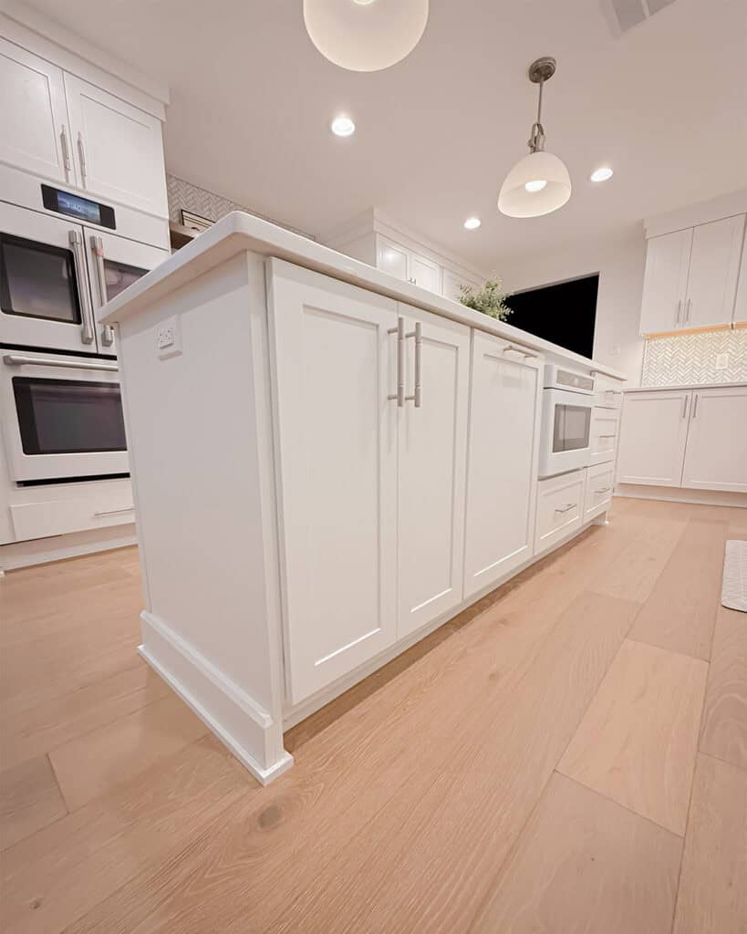 A modern kitchen with white cabinets and island, stainless steel handles, wood flooring, built-in microwave, double wall ovens, and pendant lights. The space is well-lit and clean with a contemporary design.