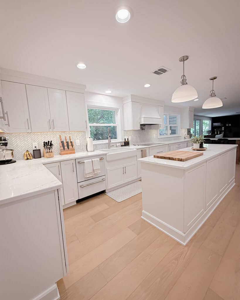 A modern kitchen with white cabinets, light wood flooring, a large island with a cutting board, farmhouse sink, stainless steel appliances, and pendant lights. The kitchen is bright and features a backsplash with a geometric pattern.