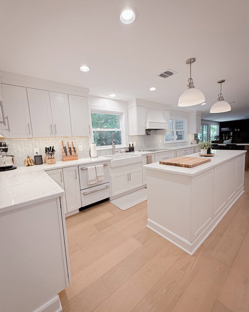 Bright, modern kitchen with white cabinets, a large island, pendant lights, light wood floors, a farmhouse sink beneath windows, and stainless steel appliances. Cutting boards and utensils sit on the counters.