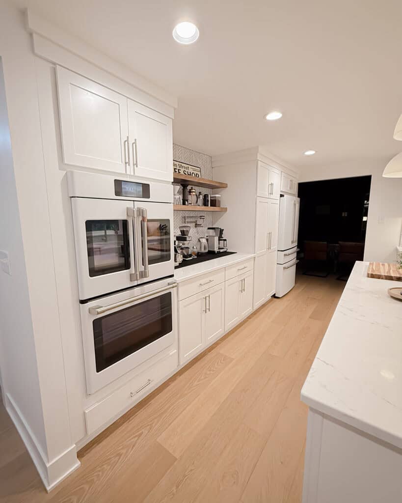 Modern kitchen with white cabinets, built-in double ovens, a coffee station, light wood flooring, and a white refrigerator. The space is well-lit with recessed ceiling lights and has a clean, minimalist design.
