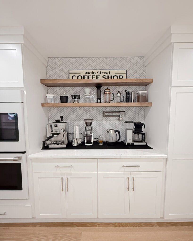 A modern home coffee bar features white cabinets, wooden shelves with mugs and jars, various coffee makers, and a Main Street Coffee Shop sign on a patterned backsplash.