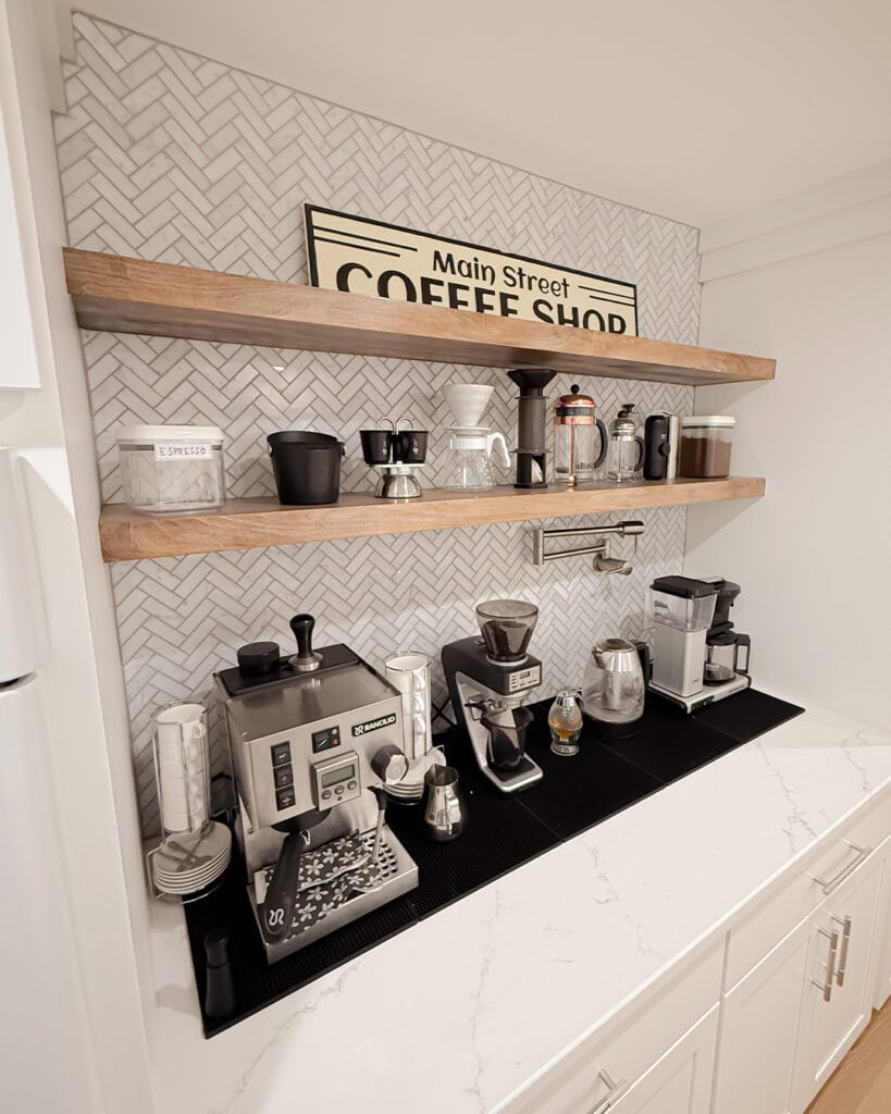 A modern home coffee bar with espresso machines, grinders, mugs, jars, and various coffee accessories neatly arranged on white counters and wooden shelves against a herringbone tile backsplash. A sign reads “Main Street Coffee Shop.”.