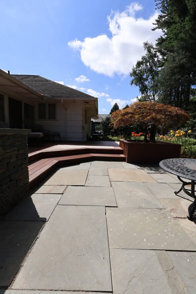 a sunny backyard patio with stone pavers, wooden steps, and railing. there is outdoor seating, a small table, a landscaped garden, and trees under a blue sky with scattered clouds.