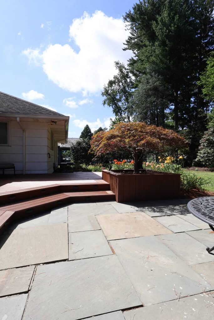 a sunlit backyard patio with stone tiles, wooden steps, a raised planter with colorful flowers, and tall trees in the background under a partly cloudy sky.