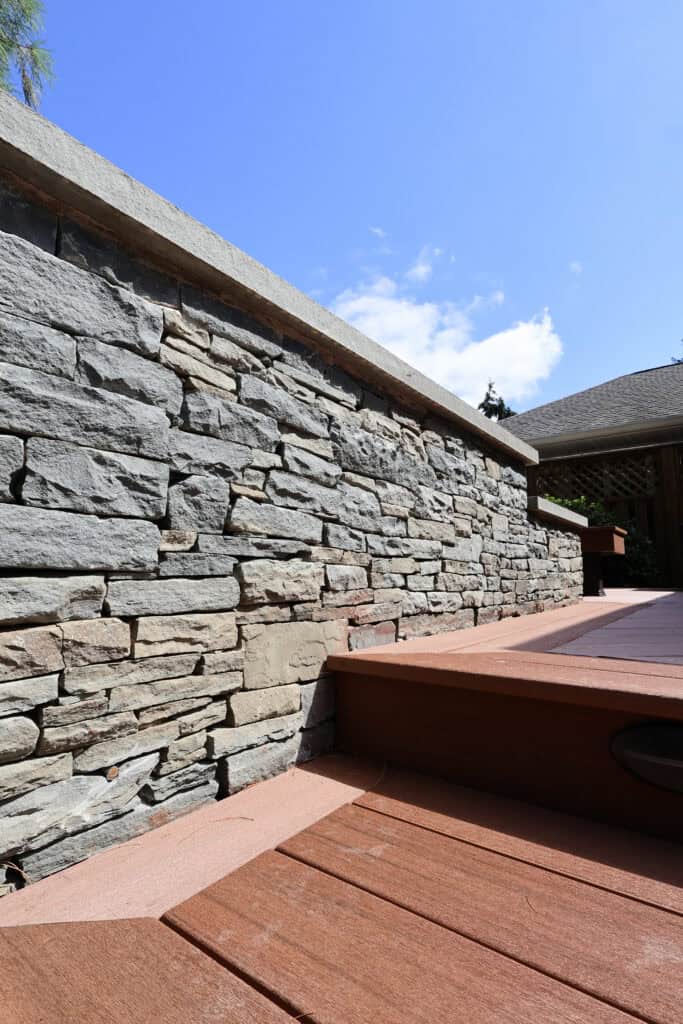 steps lead up to a wooden deck bordered by a gray stone wall, with a blue sky and a few clouds in the background.