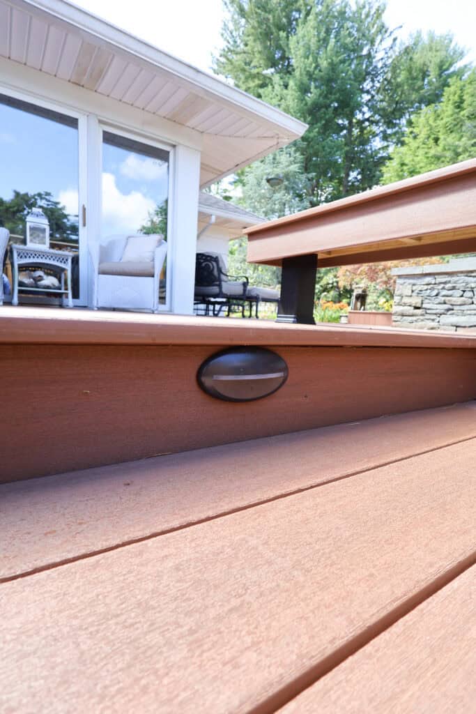 close up of a wooden deck step with a built in oval light, leading up to a patio area with outdoor furniture and glass doors. lush trees and a stone wall are visible in the background.