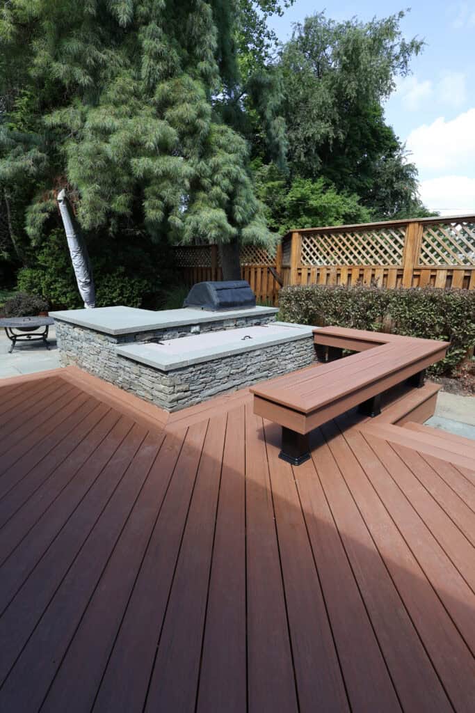 a wooden deck with built in benches surrounds a stone fire pit and grill area, fenced by wood lattice with trees and bushes in the background, on a sunny day.