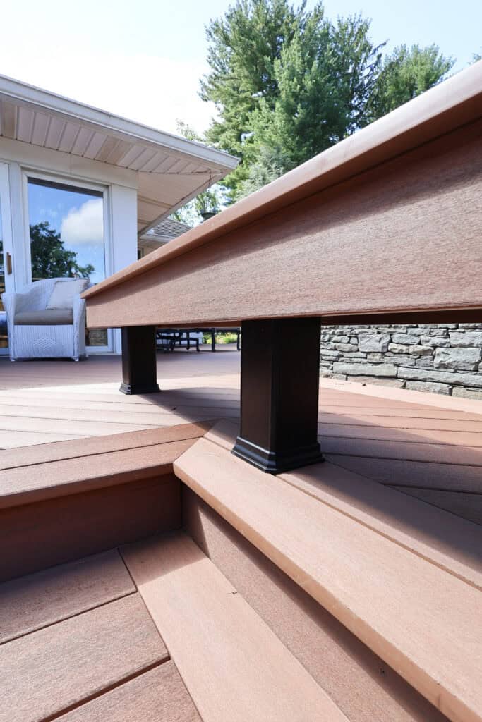 close up of brown composite deck stairs with black rail posts, leading to a patio with outdoor furniture. stone wall and trees are visible in the background under a partly cloudy sky.