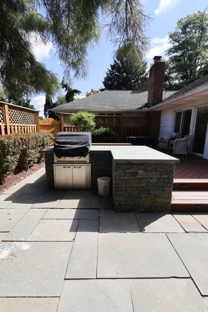 outdoor stone patio with a built in grill and covered bbq, next to a wooden deck attached to a house. trees and a wooden fence are in the background.