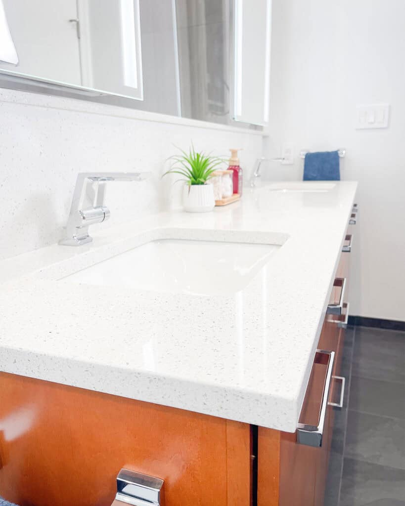 A modern bathroom countertop with two sinks, sleek chrome faucets, a potted plant, soap dispenser, and folded blue towel. The countertop is white, and the cabinets are wood with silver handles.