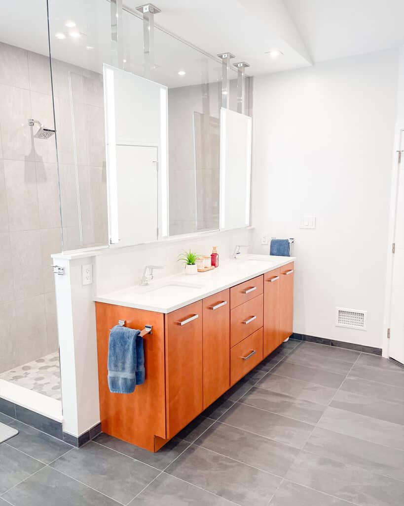 Modern bathroom with gray tile floor, large double-sink wooden vanity, two rectangular mirrors, and a glass-enclosed shower. Towels and a soap dispenser rest on the countertop, with a small potted plant for decor.