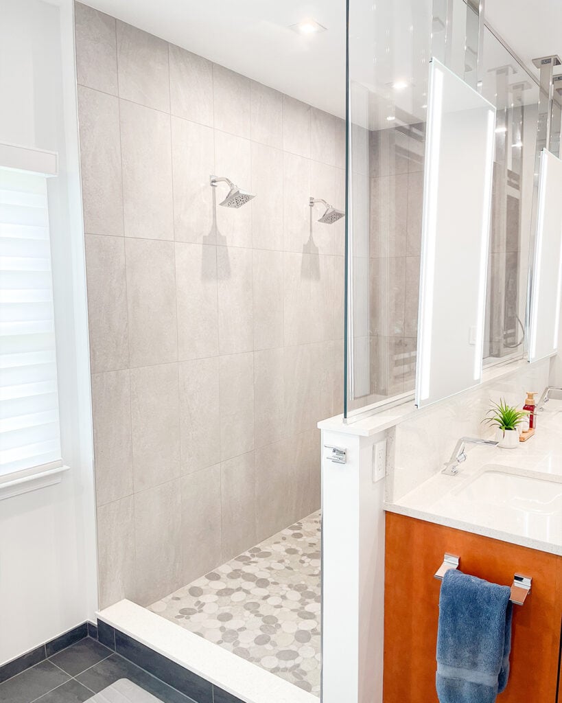 Modern bathroom with a double shower featuring two showerheads, large beige tiles on the walls, and patterned floor tiles. A white vanity with a sink, blue towel, and small potted plant is visible in the foreground.