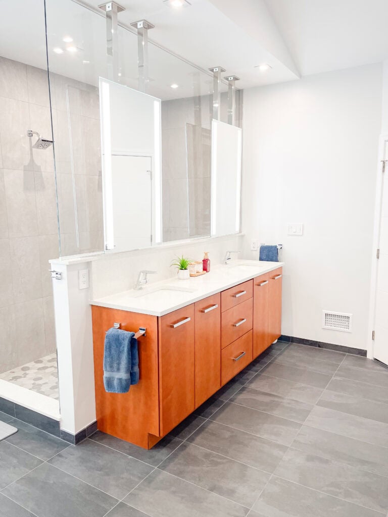 Modern bathroom with light gray tile floor, wooden double-sink vanity with white countertop, two large mirrors, towel and soap dispenser, small green plant, and a walk-in shower with glass partition.