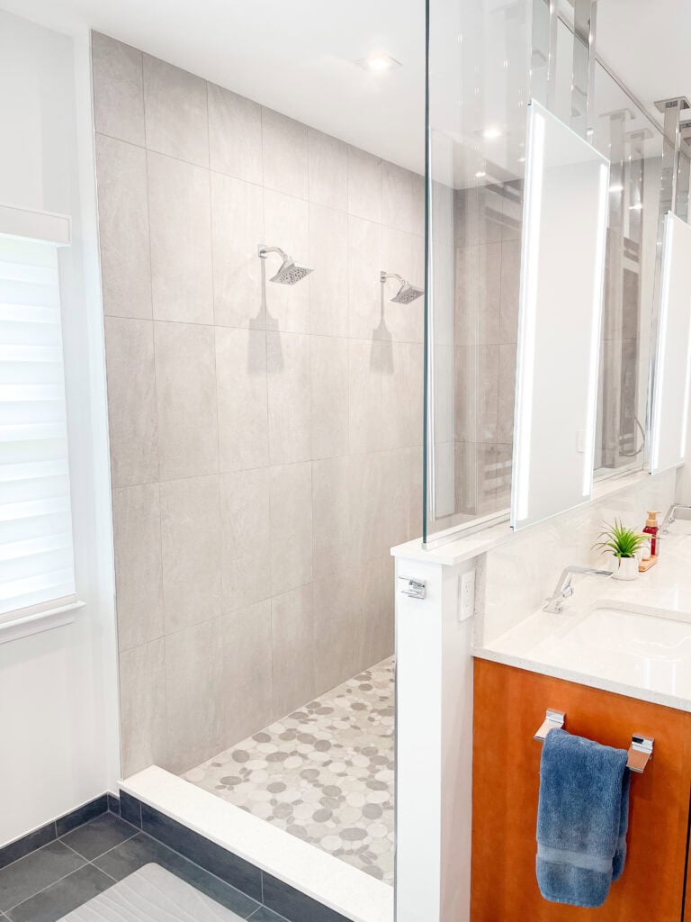 Modern bathroom with a double shower, featuring two showerheads and large gray tiles. The vanity has a white countertop, a blue towel, and a potted plant. The space is bright with natural and artificial light.