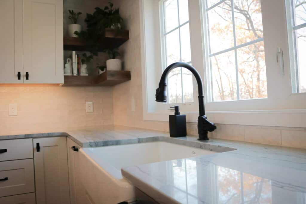 Modern kitchen with white cabinets, marble countertops, a farmhouse sink, black faucet, soap dispenser, wooden shelves with decor, and large windows letting in natural light.