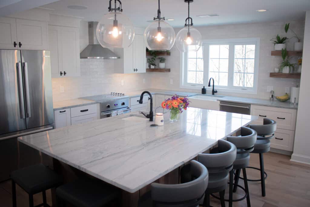 Modern kitchen with white cabinets, stainless steel appliances, marble island with gray chairs, pendant lights, flowers on the counter, and large window letting in natural light. Shelves with plants and fruit decorate the space.