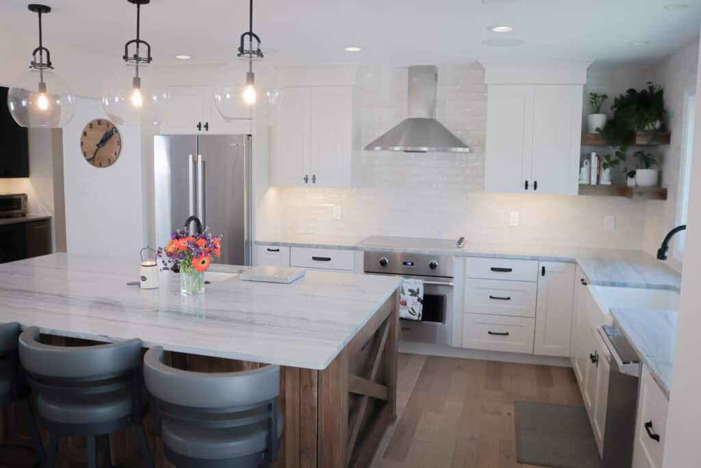 Modern kitchen with white cabinets, stainless steel appliances, a large marble island with gray chairs, pendant lights, and a vase of colorful flowers on the island. Light wood floor and shelves with plants in the corner.