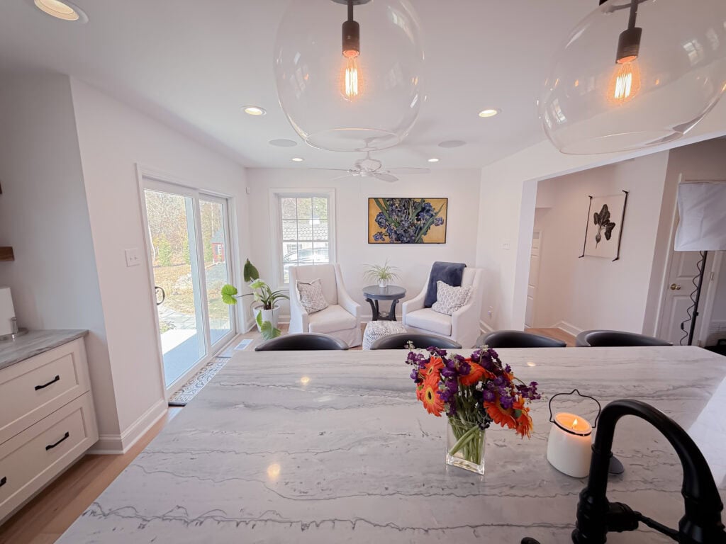 Bright, modern kitchen and living area with marble island, black chairs, and a vase of colorful flowers. Two white armchairs and plants sit near large windows, letting in natural light.