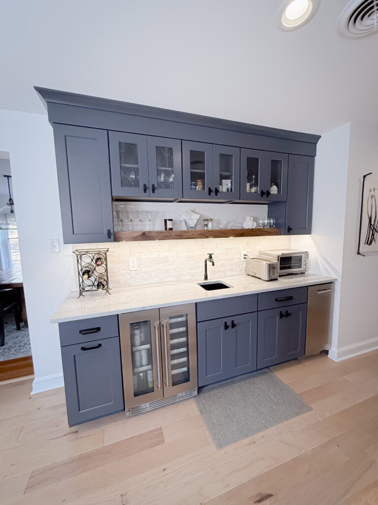 A modern kitchenette with dark blue cabinets, glass-front upper cupboards, a white tile backsplash, a small sink, a wine fridge, a microwave, and light wood flooring. A gray mat sits in front of the sink area.