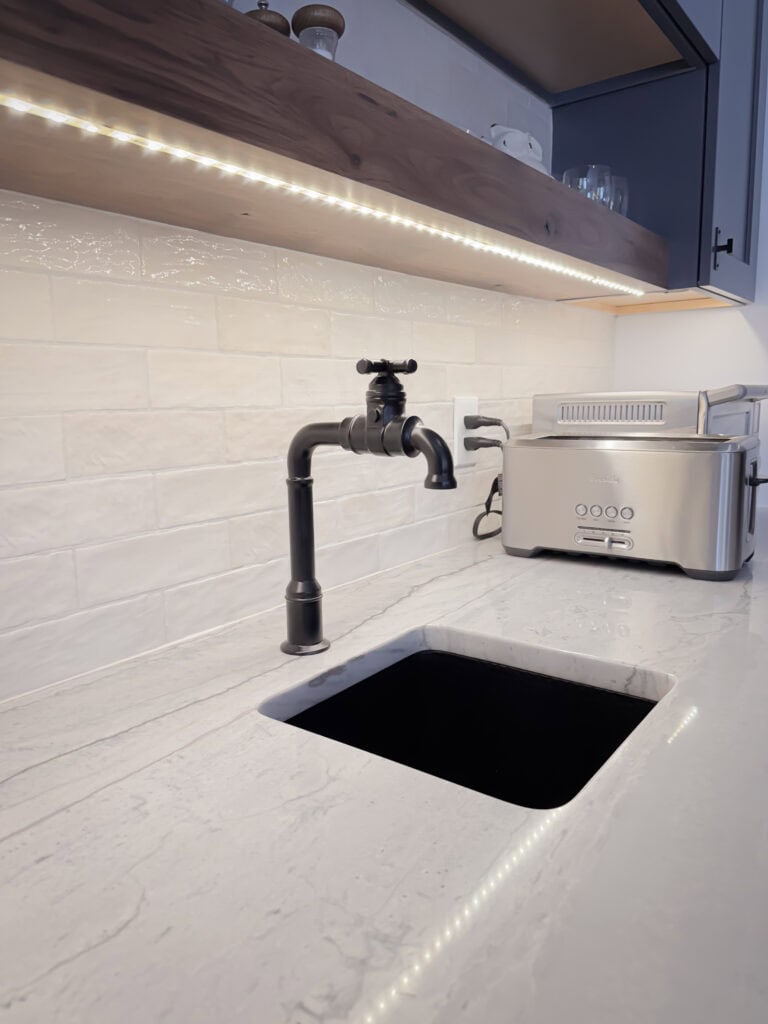 A modern kitchen counter with a black faucet, a small square sink, a stainless steel toaster, white tile backsplash, and under-shelf LED lighting.