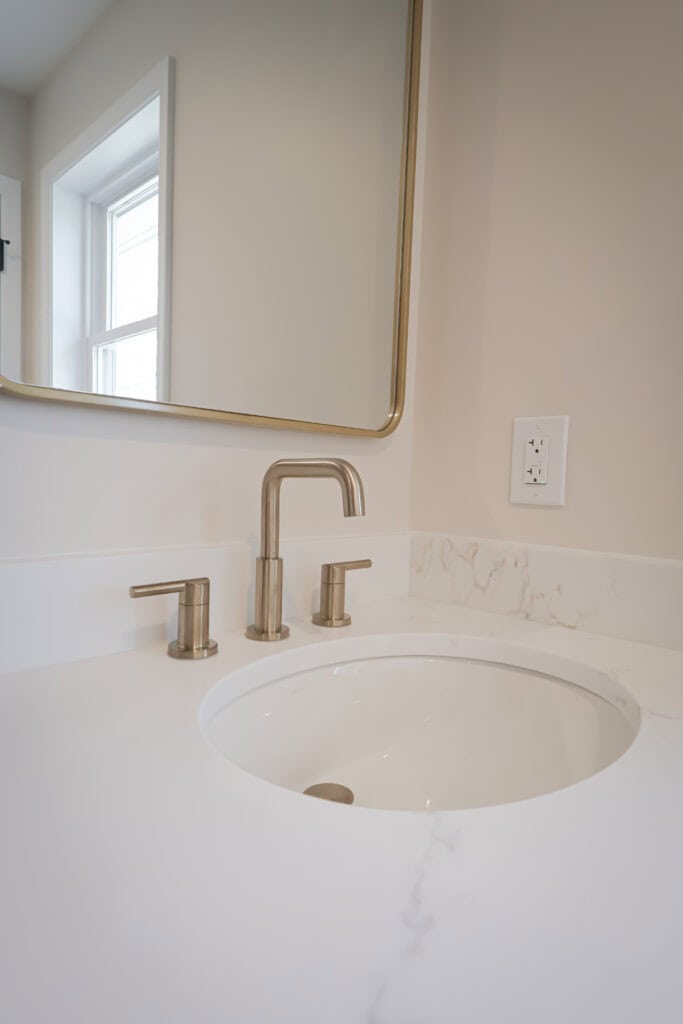 a modern bathroom sink with a marble countertop, brushed nickel faucet, and a large mirror above. a window reflects in the mirror, and an electrical outlet is on the wall beside the sink.