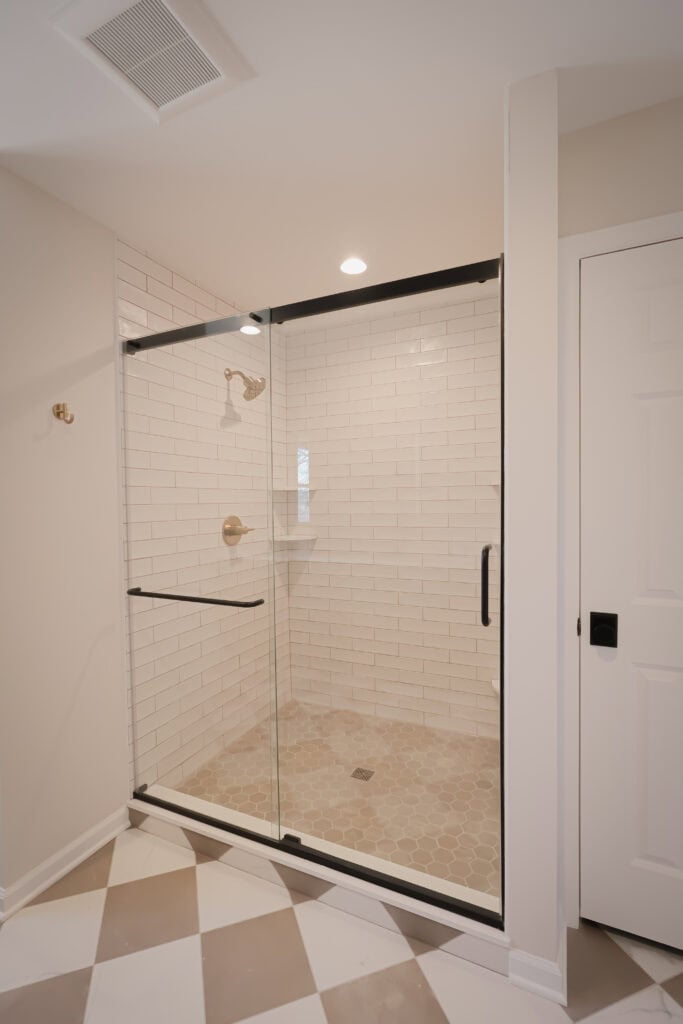 glass enclosed shower with white subway tile walls, a built in corner shelf, beige hexagon tile floor, and black hardware. the bathroom has a patterned tile floor and white walls.