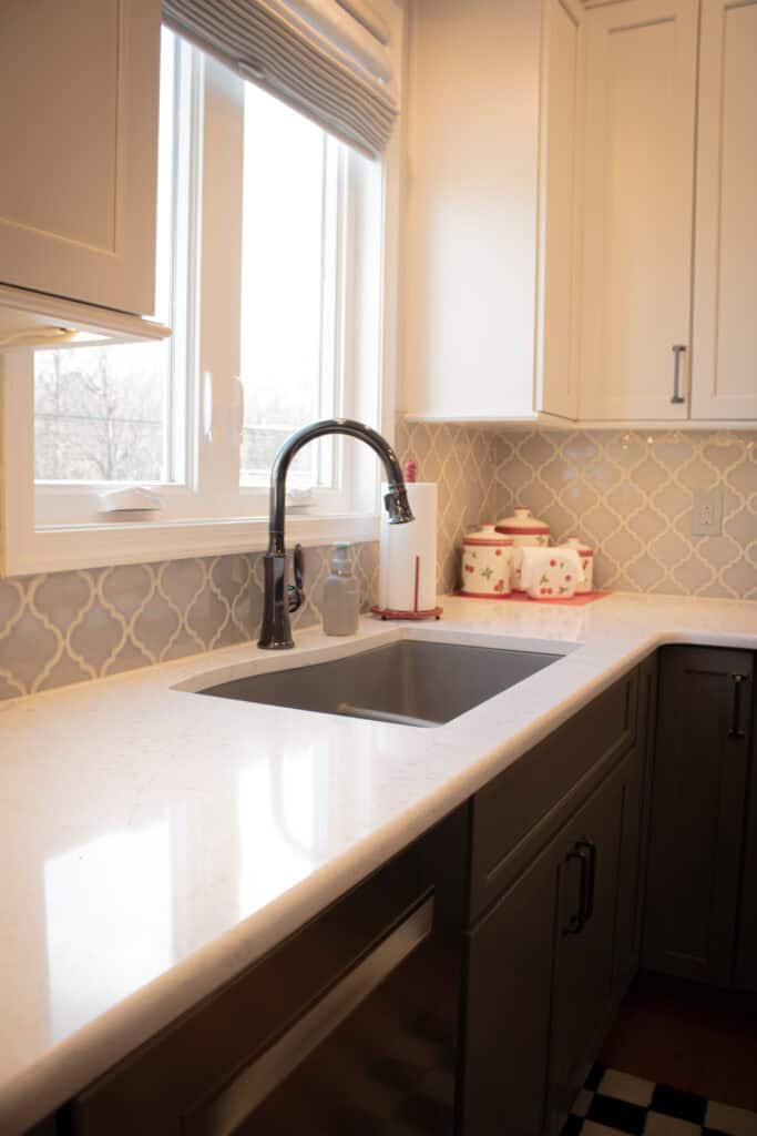 A modern kitchen sink with a tall black faucet under a large window. The countertop is white with gray cabinets below, and ceramic canisters with floral patterns sit in the corner next to a paper towel holder.