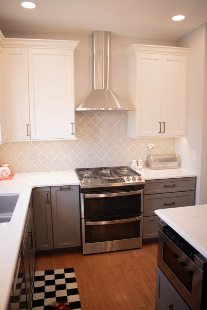 Modern kitchen with white upper cabinets, gray lower cabinets, stainless steel oven and hood, light-colored patterned backsplash, and wood flooring. Countertops are white with a breadbox and small kitchen items visible.
