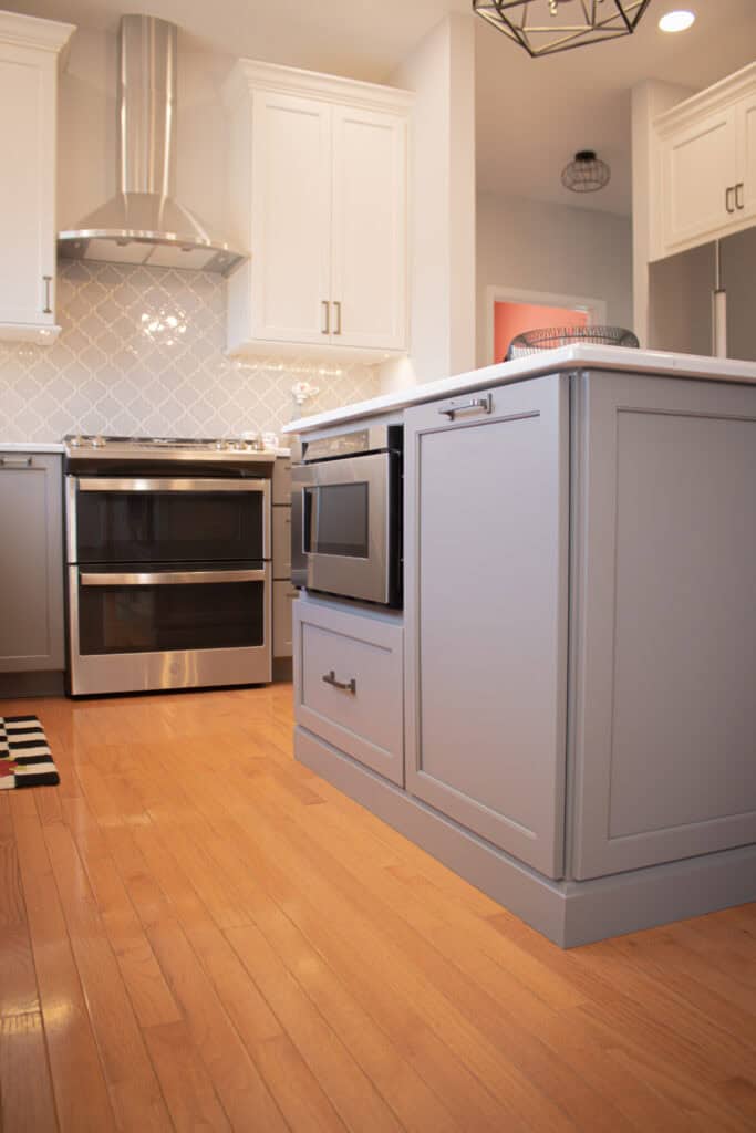 A modern kitchen with light wood flooring, gray island cabinets, a built-in microwave, stainless steel double oven, white upper cabinets, and a geometric tile backsplash.