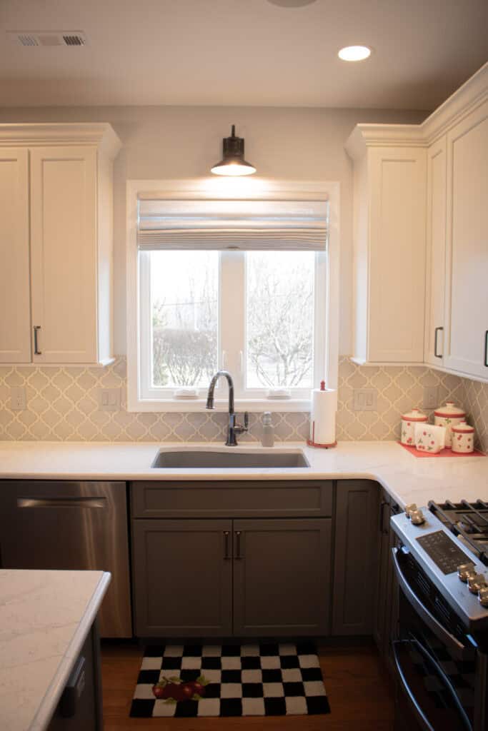 Modern kitchen with white upper cabinets, gray lower cabinets, a checkered black-and-white rug, a window above the sink, stainless steel appliances, and decorative containers on the countertop.